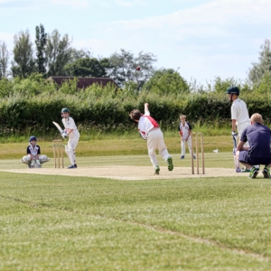 Children playing cricket on a grassy field at one of the Top 100 Cricket Schools in the UK. A bowler in action delivers a ball towards the batsman, poised for a shot. Fielders and a wicketkeeper are ready, with a coach observing from the side, surrounded by trees and bushes in the background.